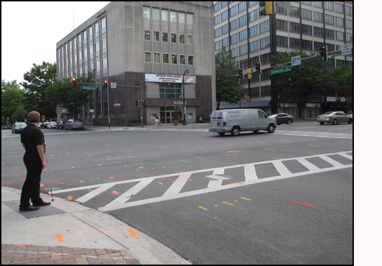Photo shows a man standing on a corner facing a 4-lane street.  To his left is a 6-lane street with a vehicle just finished crossing the intersection.
